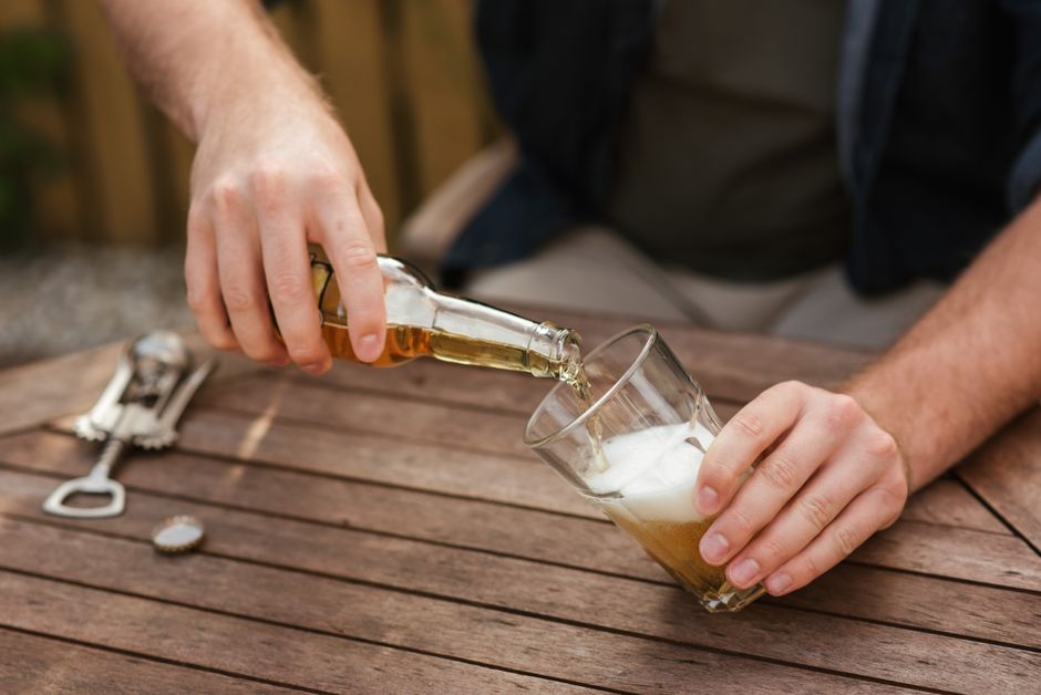From above of crop anonymous male sitting at wooden table and filling glass with beer