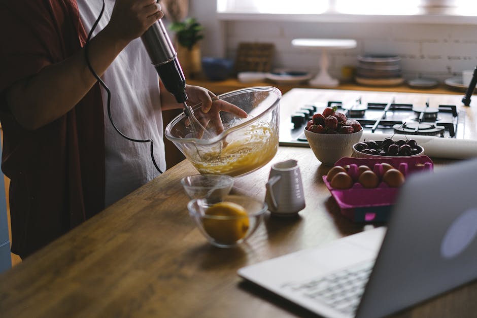 A woman uses an electric mixer to whip eggs in a cozy kitchen setting, surrounded by fruits and a laptop.