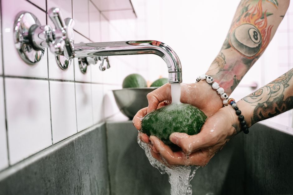 Tattooed hands washing a fresh avocado under running faucet water in a modern kitchen.