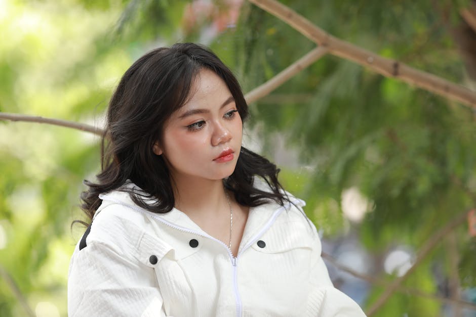A contemplative young woman in a white jacket against a lush green background.