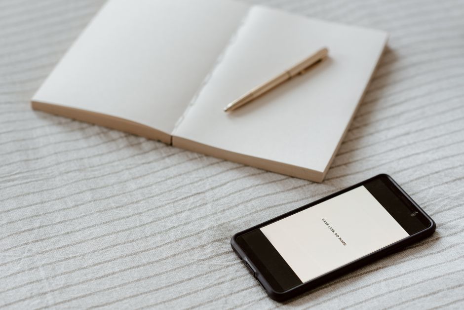 From above of cellphone with inscription on screen near blank notepad with pen on soft bed sheet