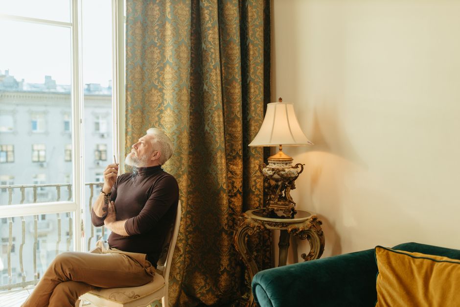A senior man with a beard reflects while sitting indoors, elegantly framed by ornate curtains and a vintage lamp.