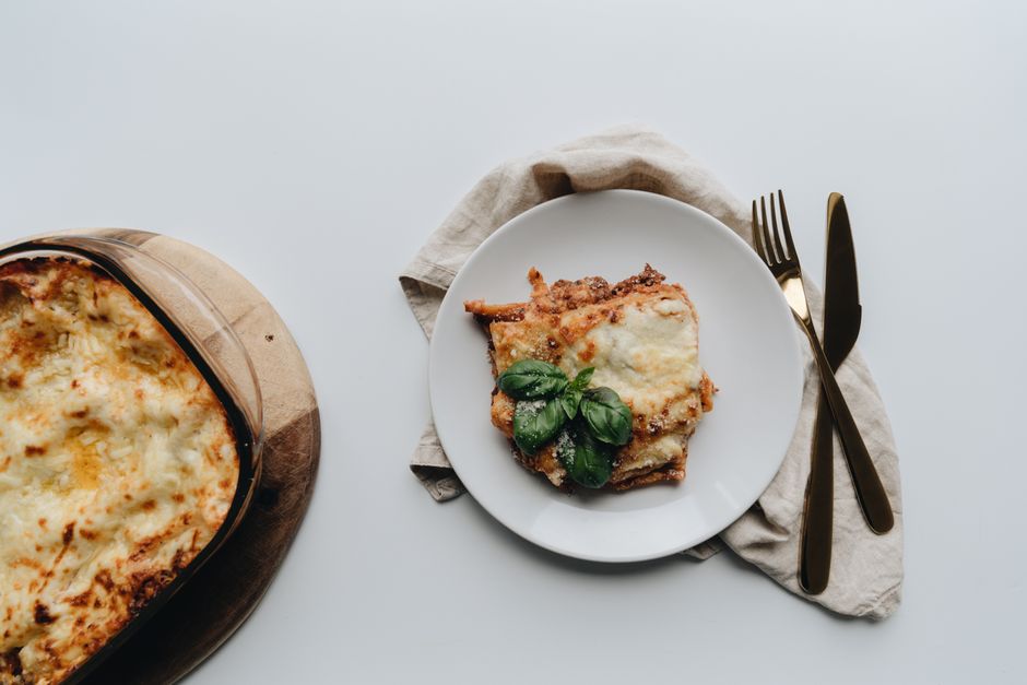 A tasty piece of homemade lasagna with basil garnish, served with cutlery on a white surface.