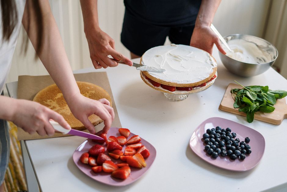 Two people preparing a homemade berry cake with fresh strawberries and blueberries.
