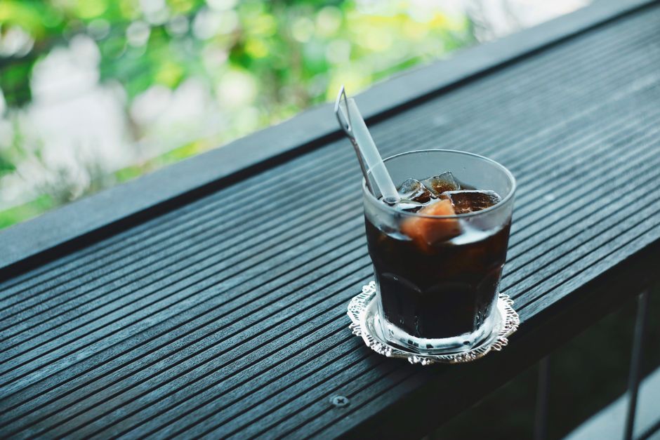A glass of iced coffee with a straw on a wooden deck amidst greenery, perfect for a summer day.